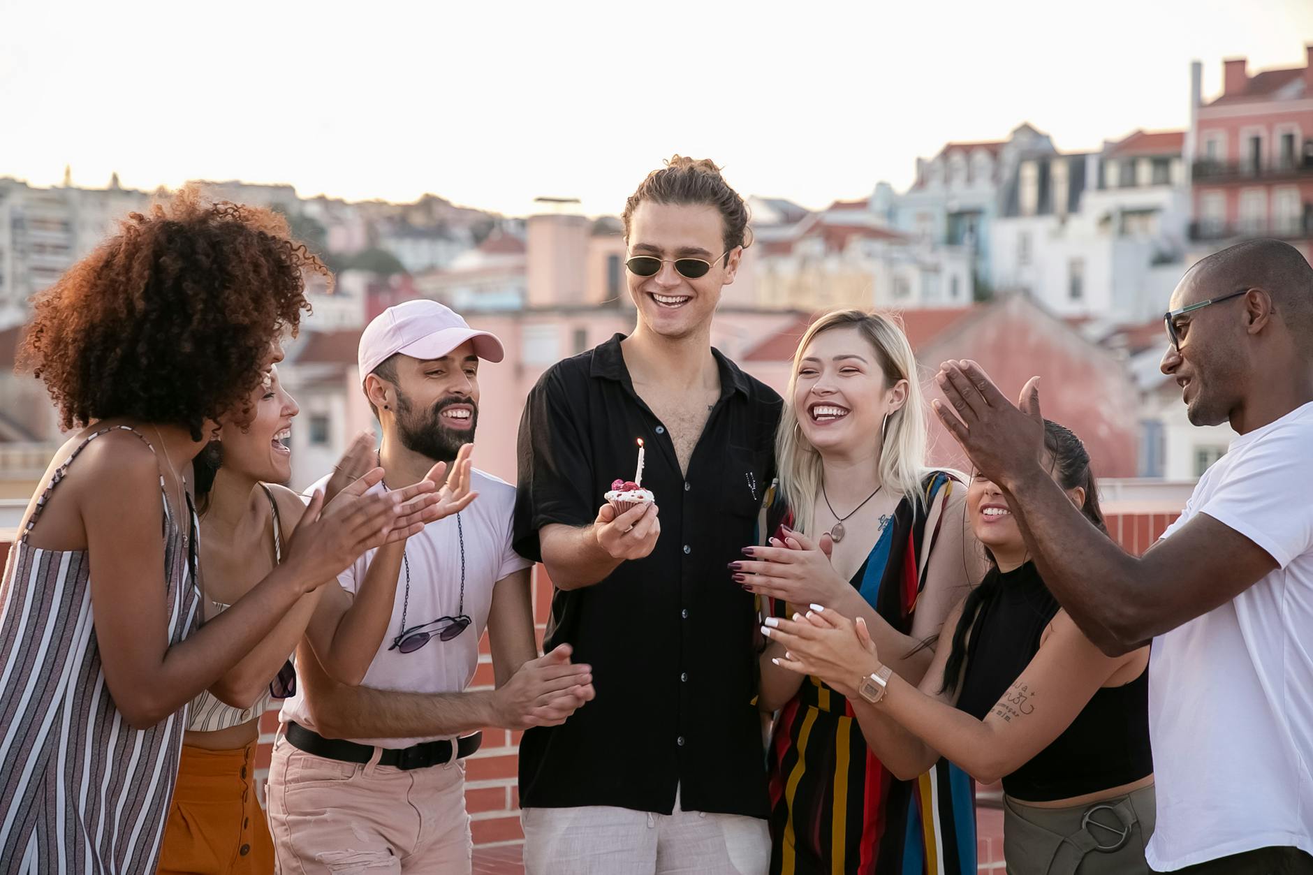 Group of young adult friends laughing and celebrating a birthday on a rooftop, holding cupcakes