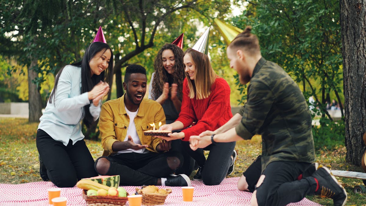 Group of teenage friends celebrating a birthday outdoors with cake and party hats
