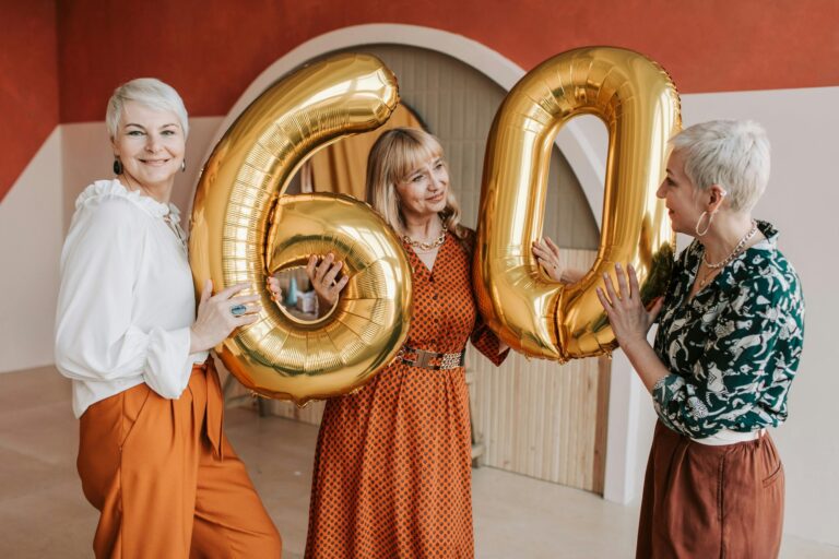 Two women in their sixties holding gold number balloons and laughing at a 60th birthday party celebration