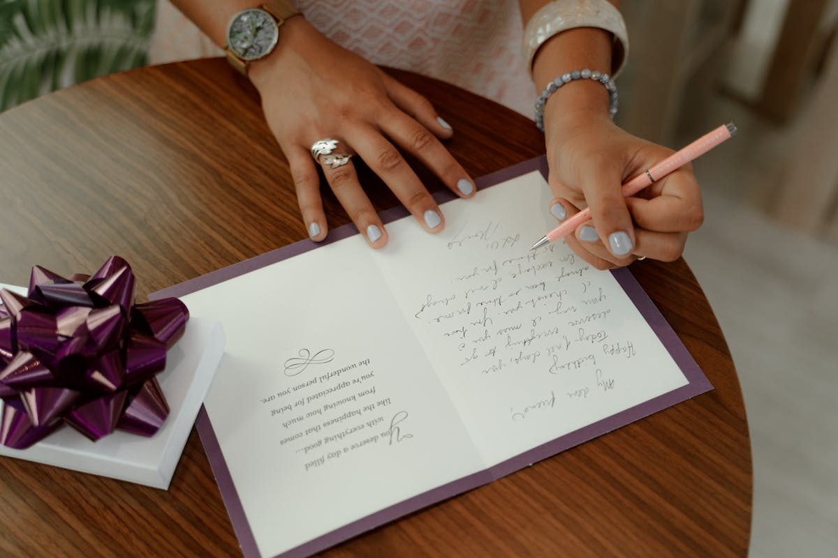 Woman writing a heartfelt birthday message inside a greeting card, with a gift box nearby