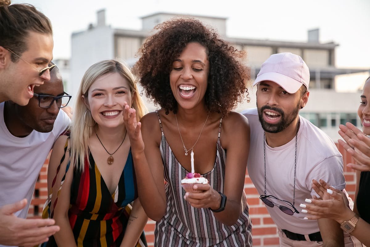 Young woman laughing while blowing out a candle on a birthday cupcake surrounded by friends outdoors