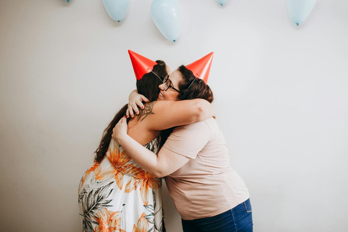 Two women hugging at a birthday party with balloons and party hats, celebrating together as sisters